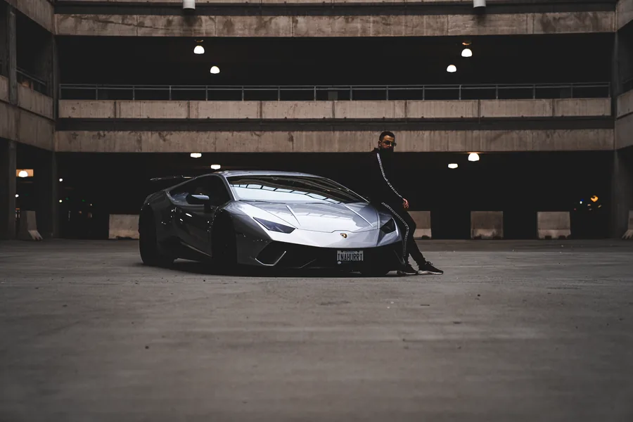 A man sitting on the bumper of a silver Lamborghini Huracán in an empty concrete parking garage