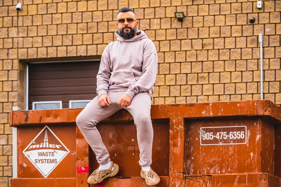 A man in a pale pink tracksuit and sunglasses sitting on an orange waste bin against a tan brick wall