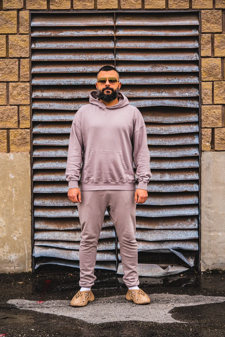 A man in a pale pink tracksuit standing in front of a large metal vent in a brick wall
