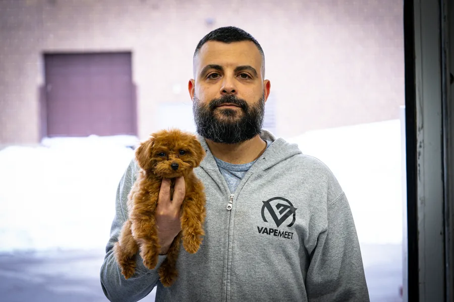 A bearded man in a grey VapeMeet zip hoodie holding a small red poodle
