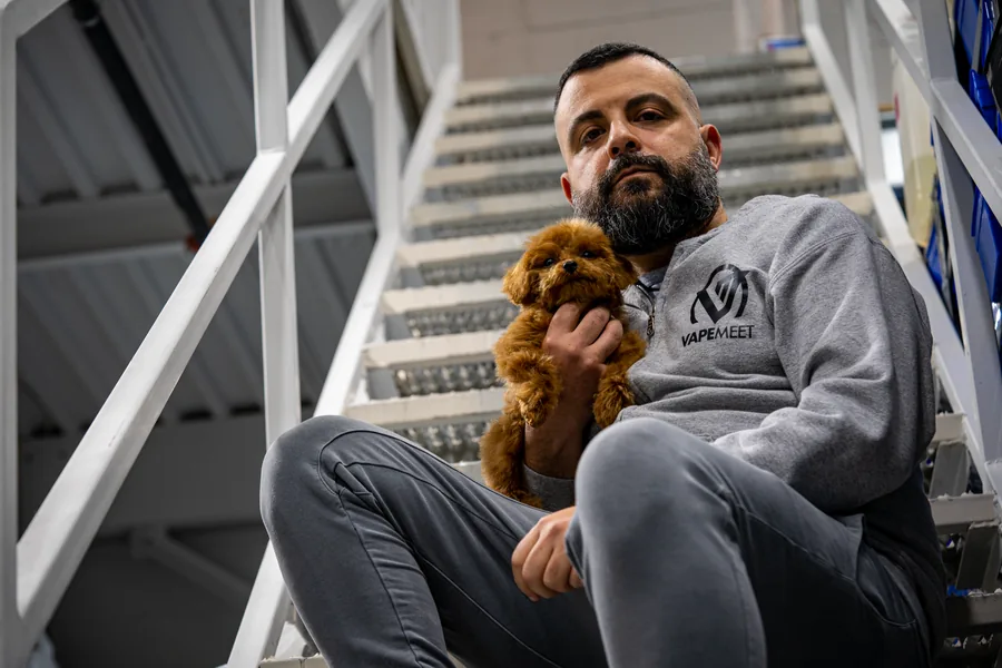 A bearded man in a grey VapeMeet hoodie sitting on white metal stairs with a small red poodle in his arm