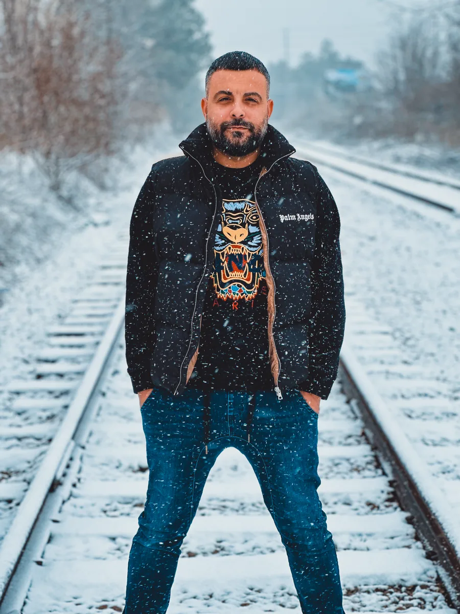 A man in a dark jacket and jeans standing on railway tracks in falling snow