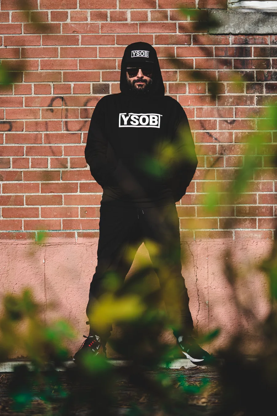 A man in a black YSOB hoodie standing against a brick wall, framed by leaves in the foreground