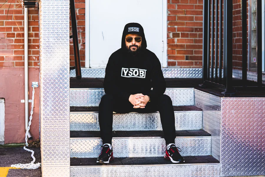 A man in a black YSOB hoodie and sunglasses sitting on diamond plate steps outside a brick building
