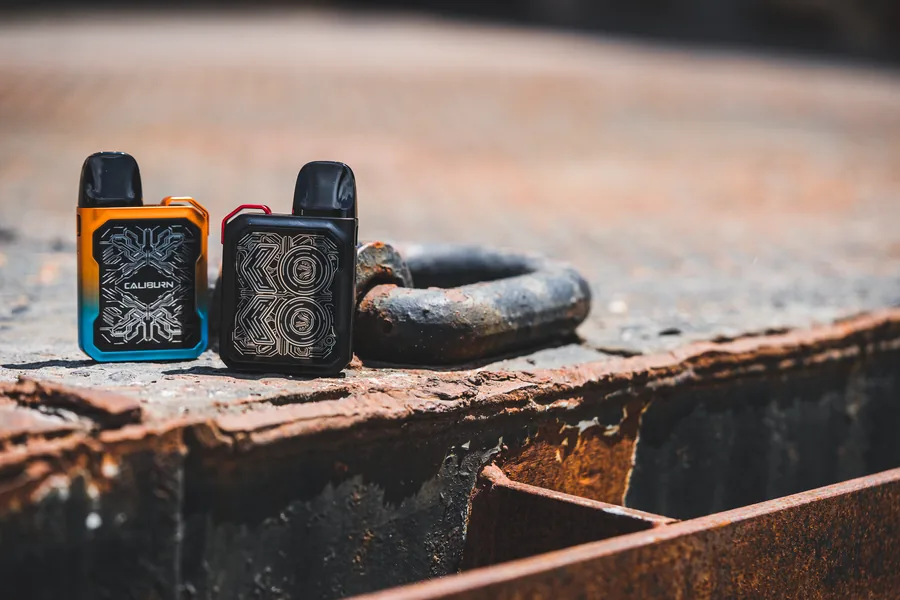 Two Uwell Caliburn pod devices with patterned fronts next to a heavy steel shackle on a rusted surface
