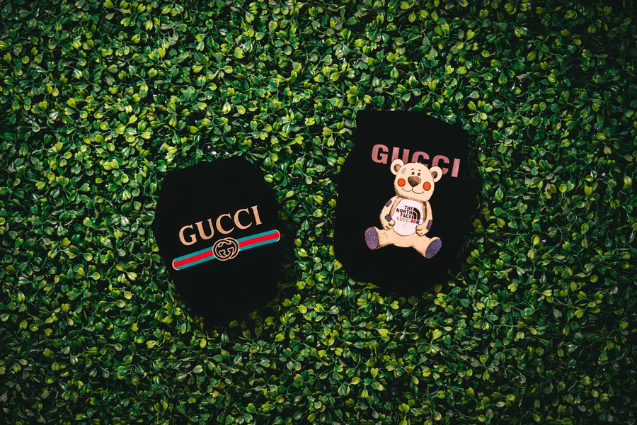 Two black Gucci patches, one with a teddy bear, laid on a green artificial hedge