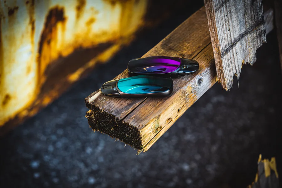 Two pod vape devices with purple and teal iridescent finishes resting on a splintered wooden beam