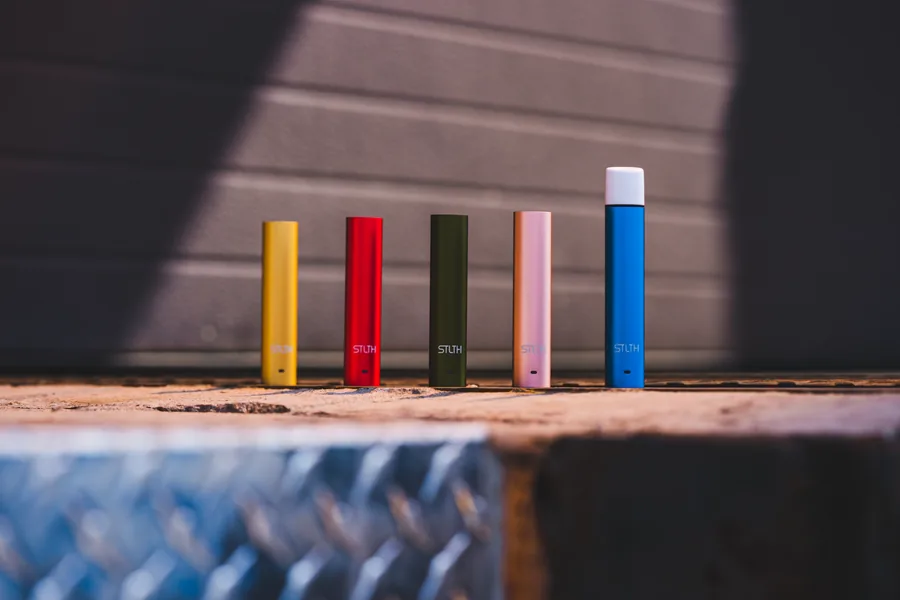 Five STLTH vape batteries in yellow, red, olive, pink and blue standing in a row on a ledge