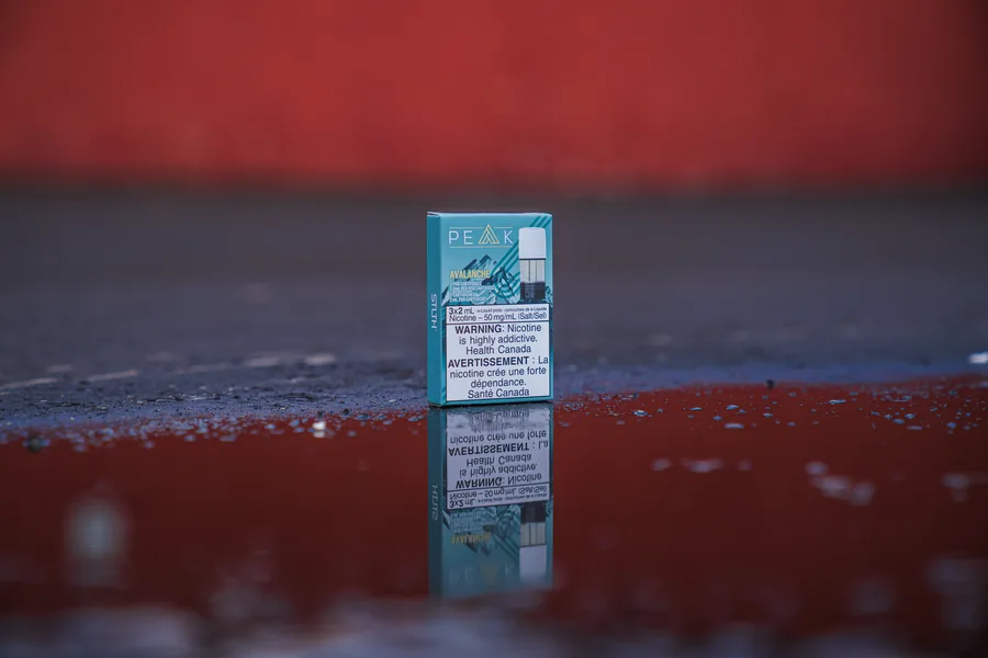 A teal STLTH Peak Avalanche pod pack standing on wet red ground with its reflection below