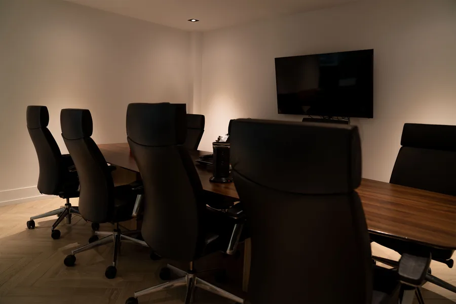 Boardroom with a long wooden table, black leather chairs and a wall mounted screen under warm lighting
