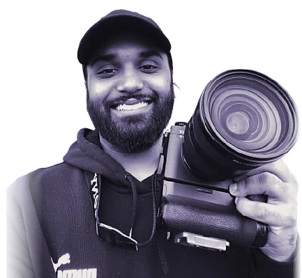 Nickolas Taki smiling in a black cap and hoodie, holding a camera up beside his face