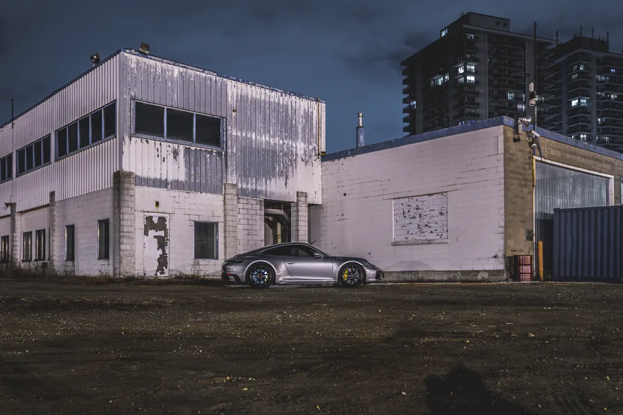 A silver Porsche 911 small in the frame in front of a weathered white industrial building under a dark sky