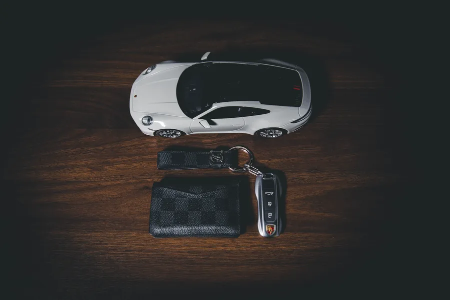 A white Porsche 911 scale model above a leather card holder and car key on a dark wooden table