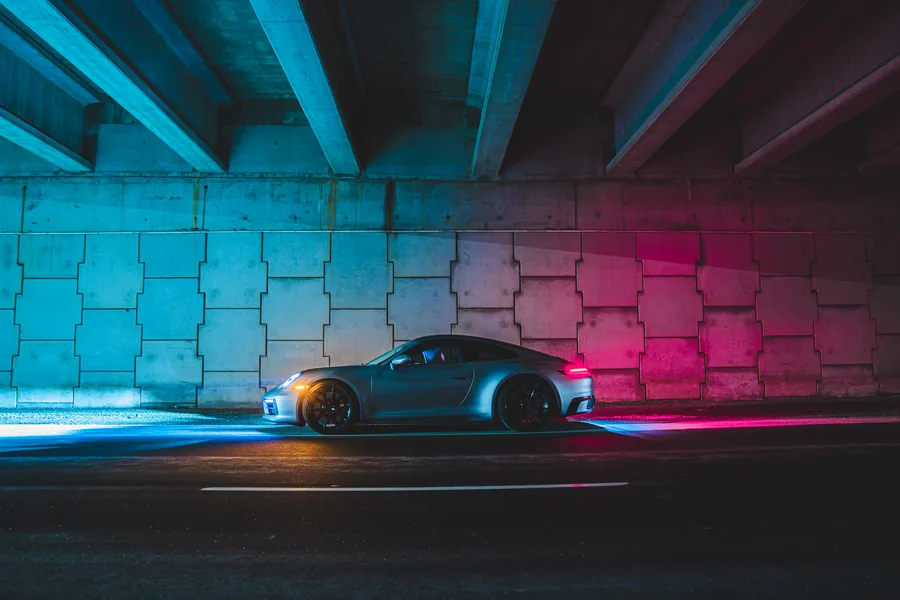 A silver Porsche 911 under a highway overpass lit in cyan and magenta
