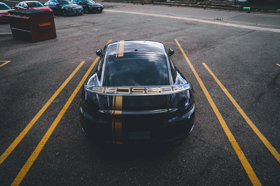 A black Porsche Cayman with gold stripes seen from above in a parking lot between yellow lines