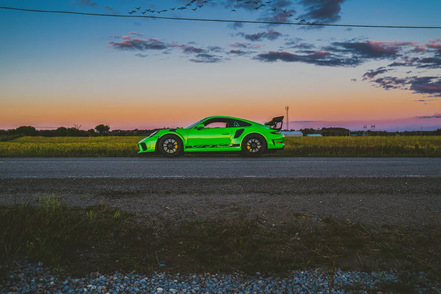 A bright green Porsche 911 with a large rear wing in profile against an orange and blue sunset