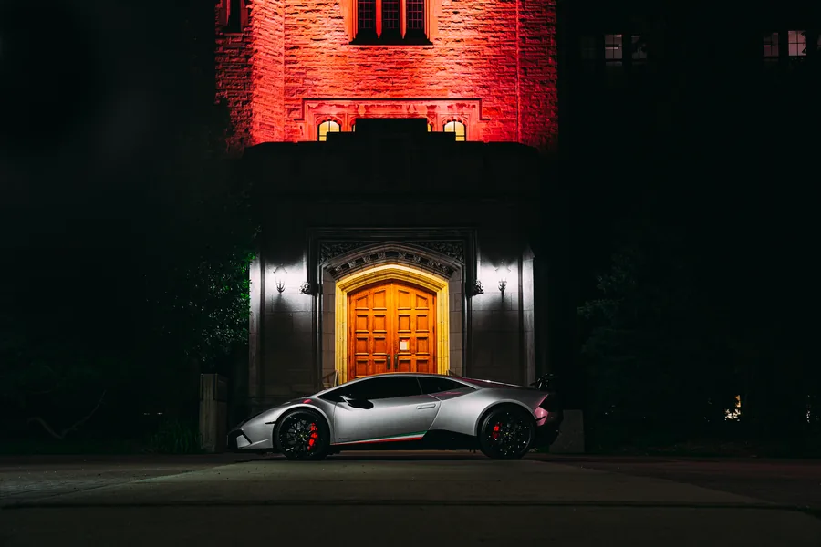 A silver Lamborghini Huracán parked at night in front of a stone church with a glowing wooden door and red lit tower