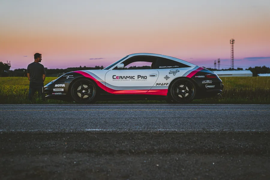 A Porsche in white, pink and black Ceramic Pro livery parked by a field at dusk with a man standing beside it