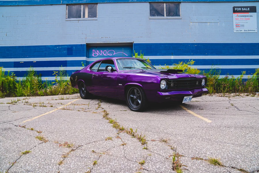 A purple classic muscle car with a black hood parked in front of a blue and white striped industrial wall